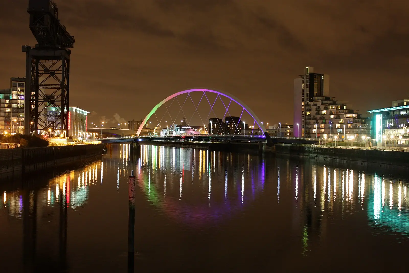 The lit Clyde Arc bridge and riverside buildings reflected in the River Clyde at night in Glasgow