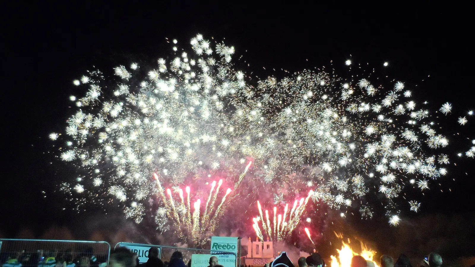 Large white and pink fireworks burst over a bonfire and a crowd at Lewes on Bonfire Night in 2013